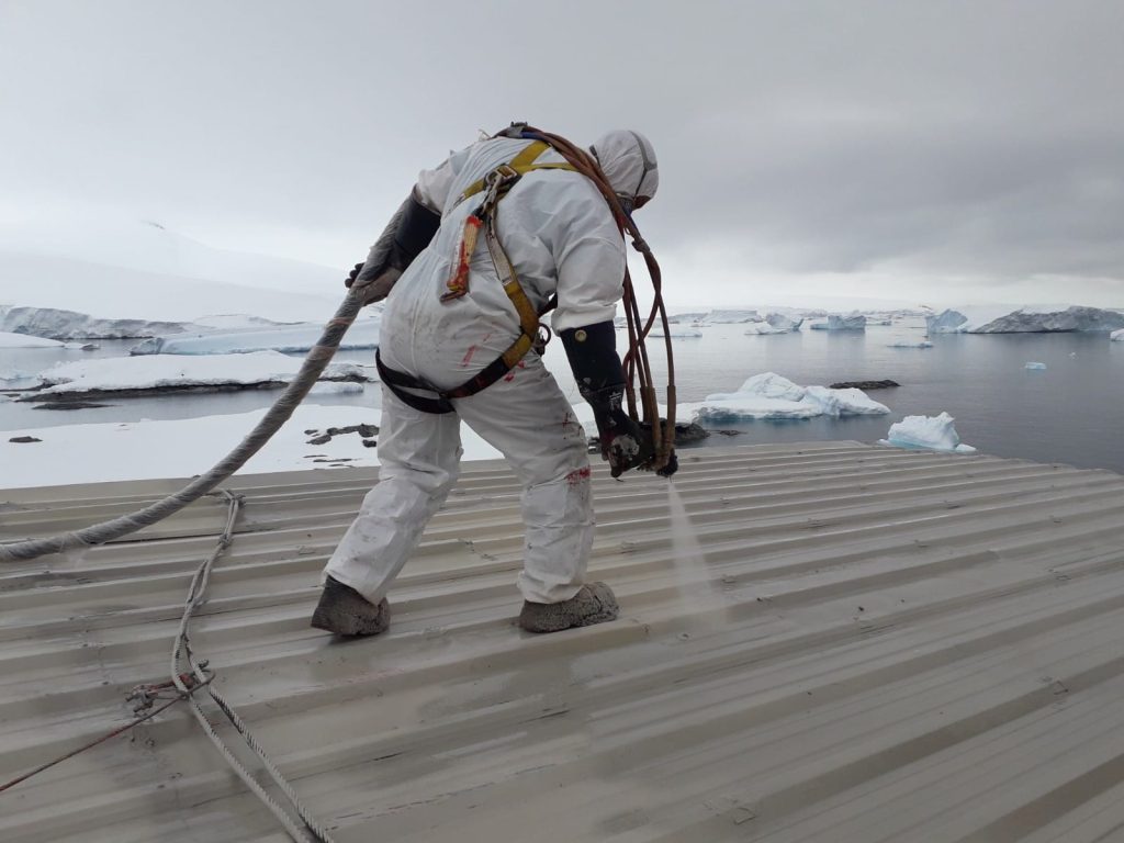 Operario de Aispoltec proyectando poliurea sobre un techo metálico en la Antártica, con icebergs de fondo